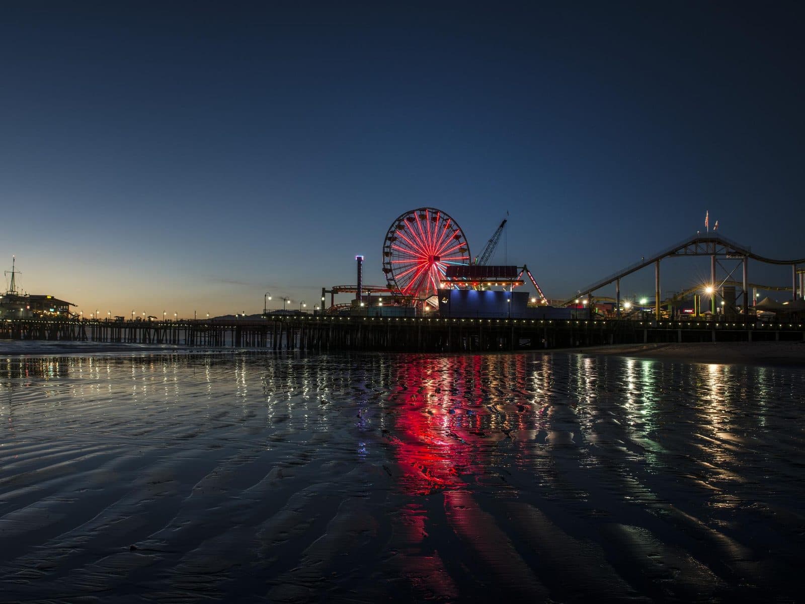 Santa Monica Pier and Pacific Park Ferris wheel lit up at sunset, reflected on the beach in Santa Monica, California